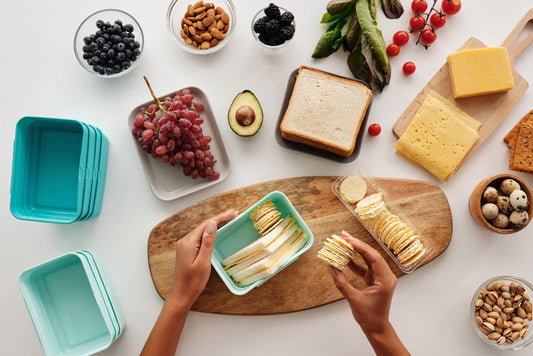 Mani che preparano un contenitore per il pranzo con pane e cracker su un tavolo con ingredienti e lunchbox